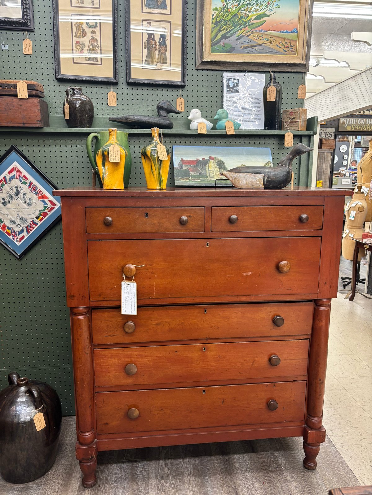 Vintage wooden dresser with multiple drawers displayed in an antique shop with pegboard wall and decorative items