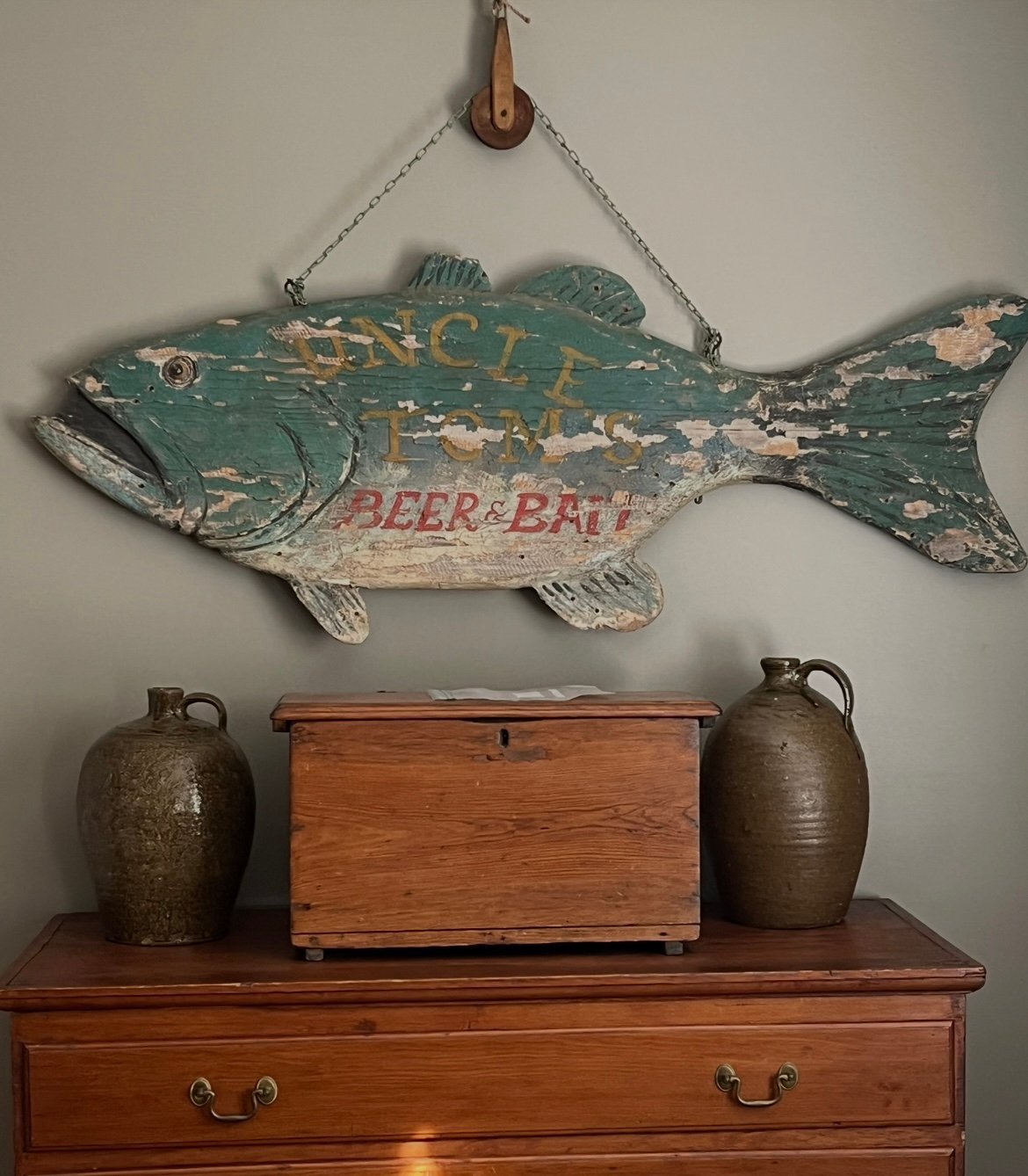 Vintage wooden fish sign painted green and cream with BEER & BAIT text, hanging above wooden furniture with ceramic jugs