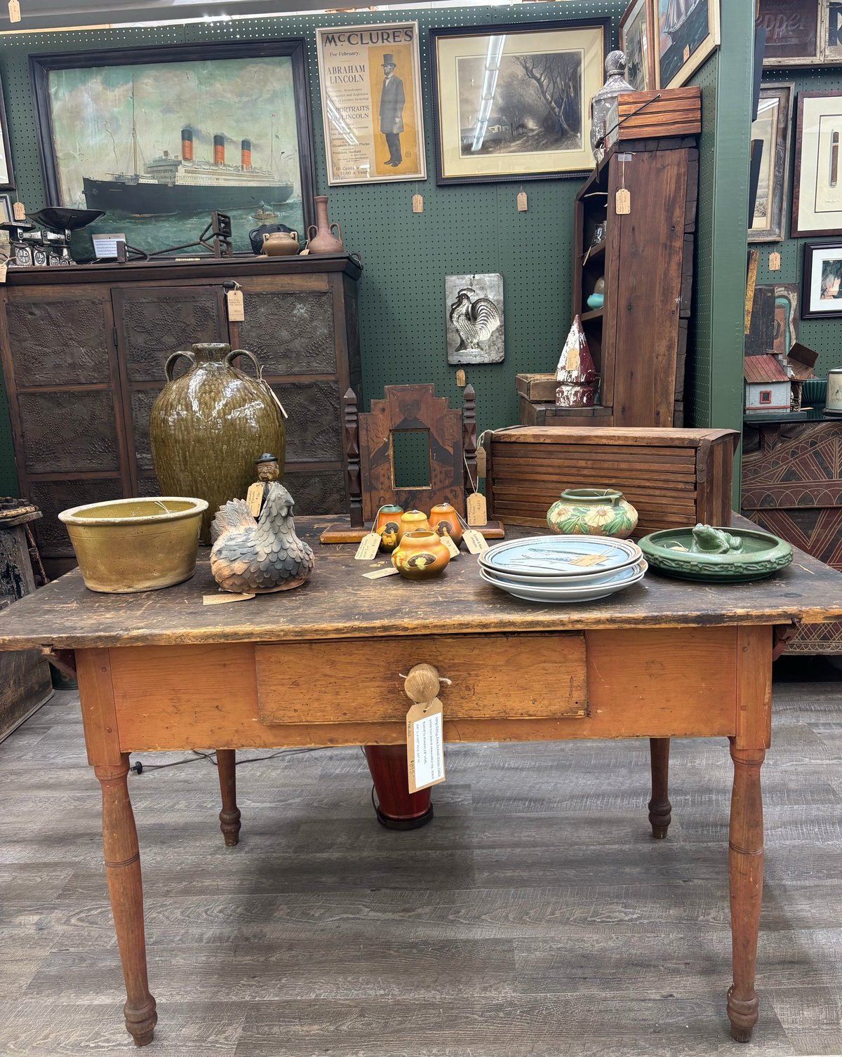 Antique wooden table with vintage kitchenware, pottery, and decorative items displayed in a green-walled shop or museum setting