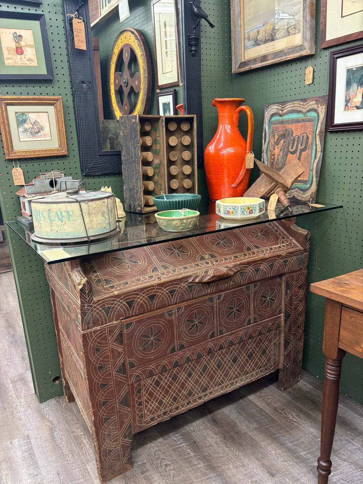 Antique wooden chest with decorative geometric patterns in a thrift store display with framed art, vintage pitcher, and collectibles on top