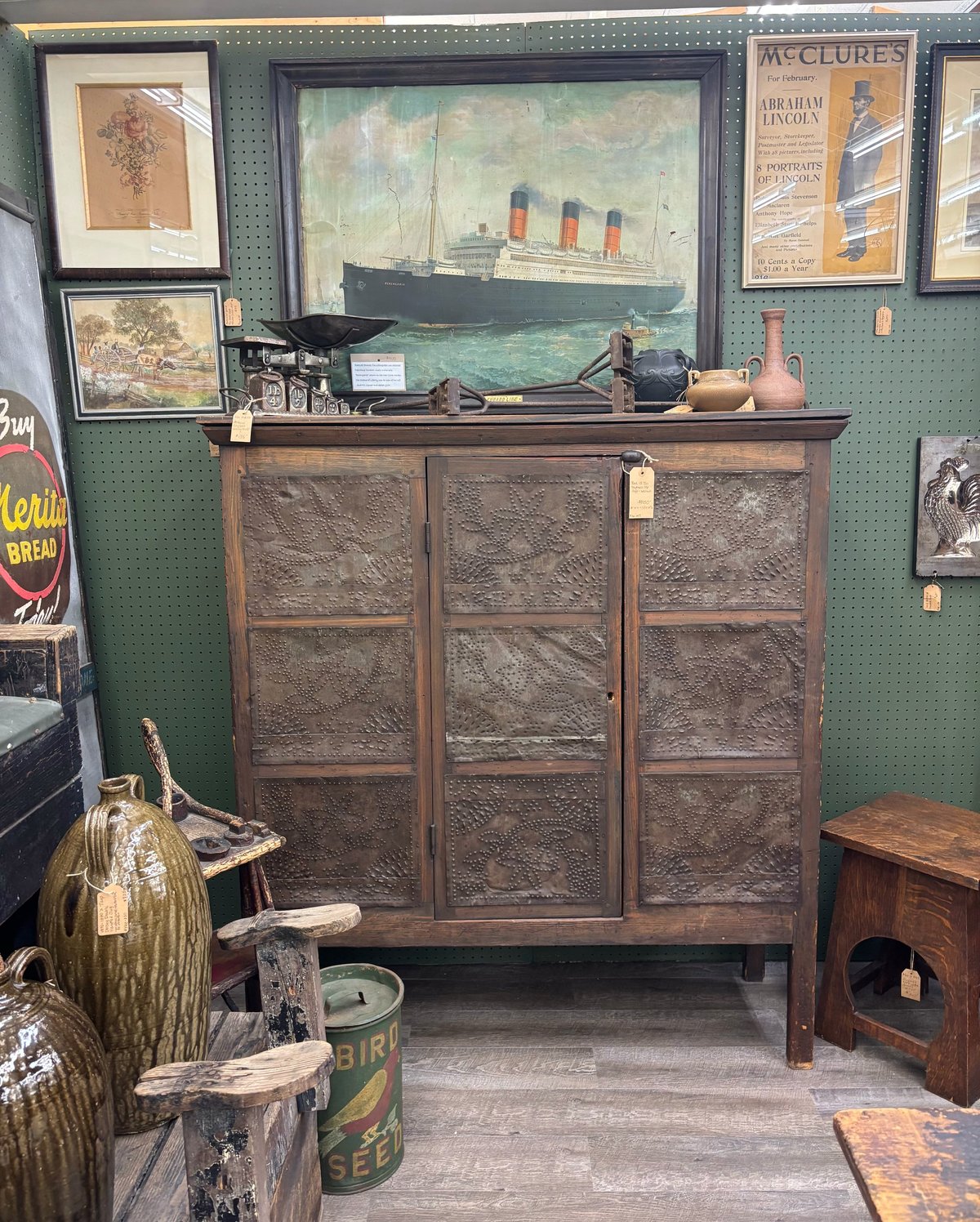 Vintage wooden cabinet with tin panels displayed in antique shop, surrounded by maritime artwork and decorative items